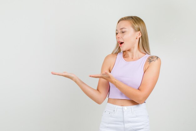 Mujer joven en camiseta, minifalda manteniendo las palmas vacías mientras mira a un lado y se ve alegre