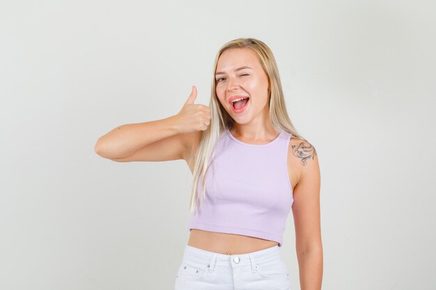 Mujer joven en camiseta, minifalda mostrando el pulgar hacia arriba con ojos parpadeados y mirando feliz