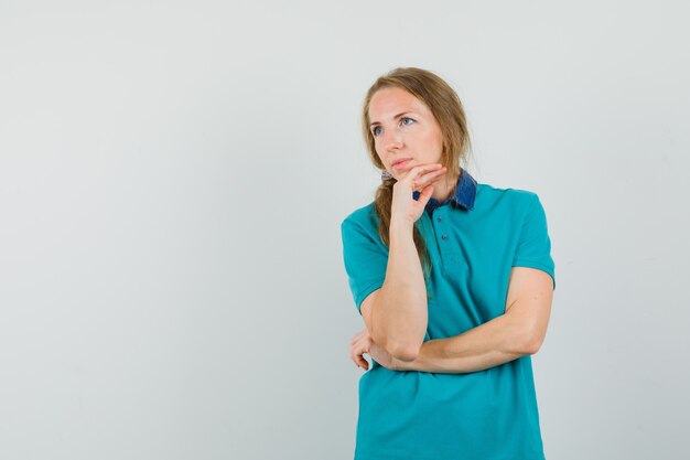 Mujer joven en camiseta mirando a un lado con la barbilla apoyada en la mano y mirando pensativo
