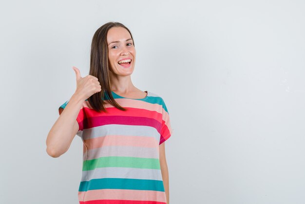 Mujer joven en camiseta mostrando el pulgar hacia arriba y mirando feliz, vista frontal.