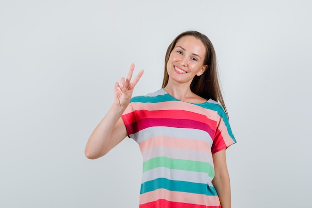 Mujer joven en camiseta mostrando el signo de la victoria y mirando alegre, vista frontal.