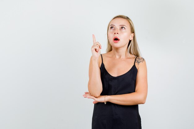Mujer joven en camiseta negra apuntando hacia arriba con el dedo y mirando enfocado