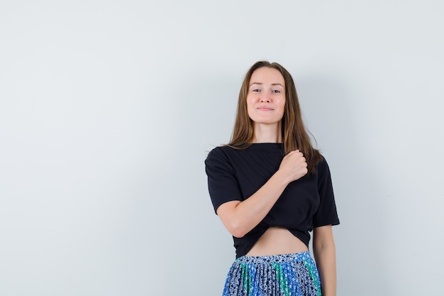 Mujer joven en camiseta negra y falda azul apretando el puño, sosteniendo la mano en el pecho y luciendo atractiva