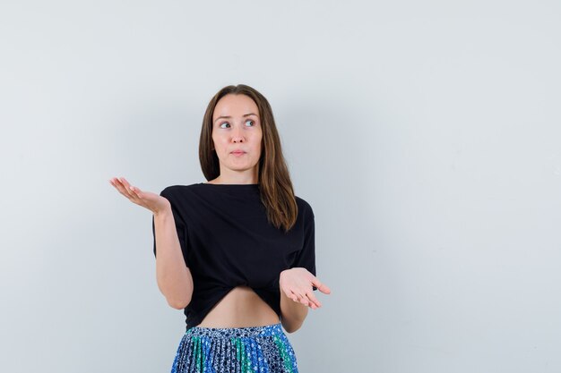 Foto gratuita mujer joven en camiseta negra y falda azul levantando una mano mientras sostiene algo mientras estira la otra mano para recibir algo y lucir atractivo