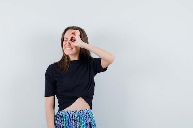 Mujer joven en camiseta negra y falda azul que muestra el signo de ok en el ojo y parece feliz