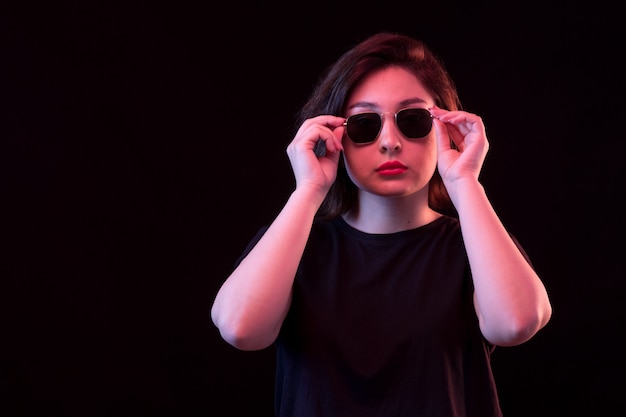 Mujer joven en camiseta negra y gafas de sol posando en la pared negra