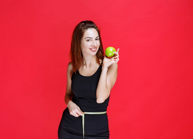 Mujer joven en camiseta negra sosteniendo manzanas verdes con una cinta métrica en la cintura