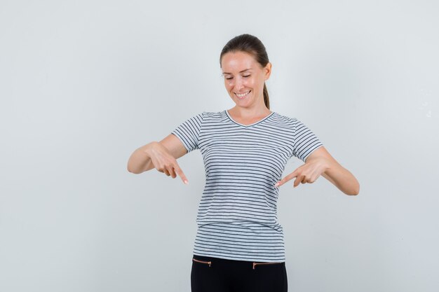 Mujer joven en camiseta, pantalones apuntando hacia abajo y mirando alegre, vista frontal.