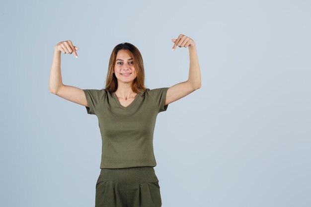 Foto gratuita mujer joven en camiseta, pantalones apuntando hacia abajo y mirando feliz, vista frontal.