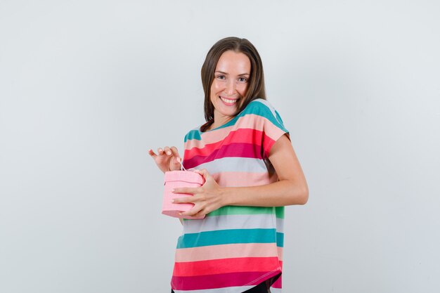 Mujer joven en camiseta, pantalones con caja de regalo y mirando contento, vista frontal.