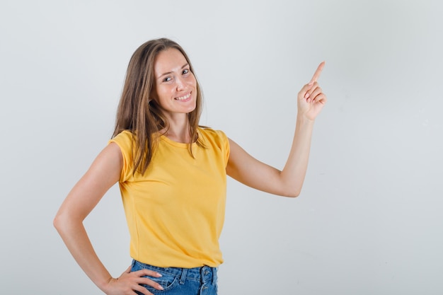 Mujer joven en camiseta, pantalones cortos apuntando con el dedo hacia arriba y mirando alegre