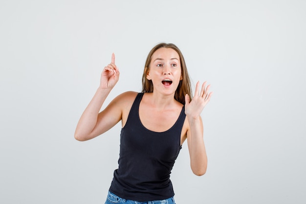 Mujer joven en camiseta, pantalones cortos apuntando con el dedo hacia arriba con signo de mano