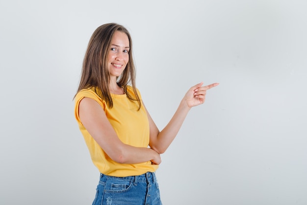 Mujer joven en camiseta, pantalones cortos apuntando con el dedo a un lado y mirando alegre