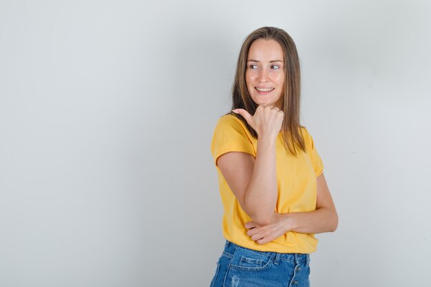 Mujer joven en camiseta, pantalones cortos apuntando hacia el lado con el pulgar y mirando alegre