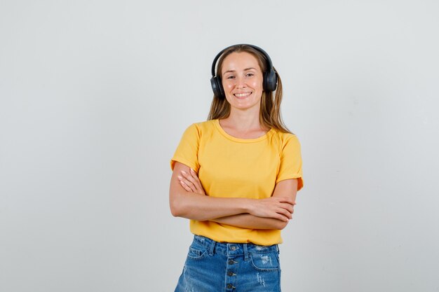 Mujer joven en camiseta, pantalones cortos, auriculares cruzando los brazos y sonriendo