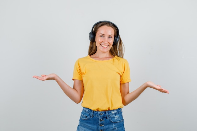 Mujer joven en camiseta, pantalones cortos, auriculares mostrando gesto de impotencia y mirando contento