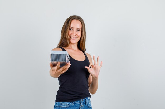 Mujer joven en camiseta, pantalones cortos con caja de regalo con signo de ok y mirando alegre