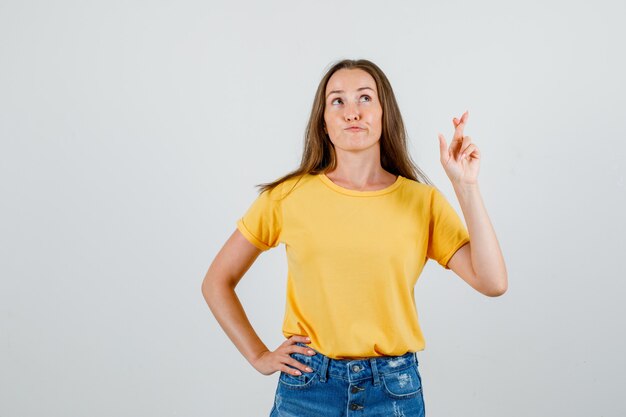 Mujer joven en camiseta, pantalones cortos cruzando los dedos mientras mira hacia arriba y parece esperanzado