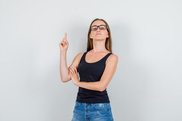 Mujer joven en camiseta, pantalones cortos, gafas apuntando hacia arriba con el dedo y mirando confiado