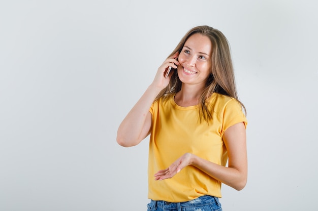 Mujer joven en camiseta, pantalones cortos hablando por teléfono inteligente y mirando alegre