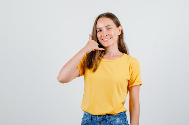 Mujer joven en camiseta, pantalones cortos mostrando gesto de teléfono y mirando feliz