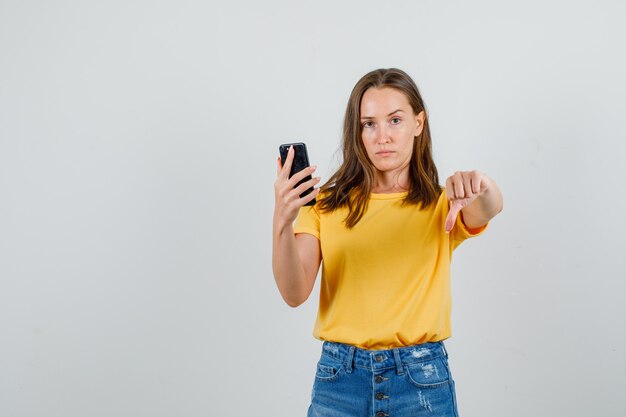 Mujer joven en camiseta, pantalones cortos mostrando el pulgar hacia abajo mientras sostiene el teléfono inteligente y parece triste