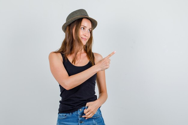 Mujer joven en camiseta, pantalones cortos, sombrero apuntando hacia un lado y mirando lindo