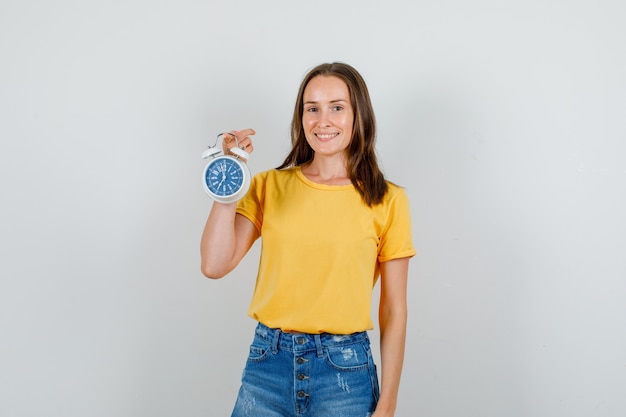 Mujer joven en camiseta, pantalones cortos sosteniendo despertador y sonriendo