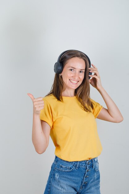 Mujer joven en camiseta, pantalones cortos tocando los auriculares con el pulgar hacia arriba y mirando feliz