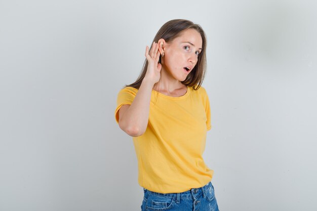 Mujer joven en camiseta, pantalones cortos tratando de escuchar el secreto y mirando enfocado