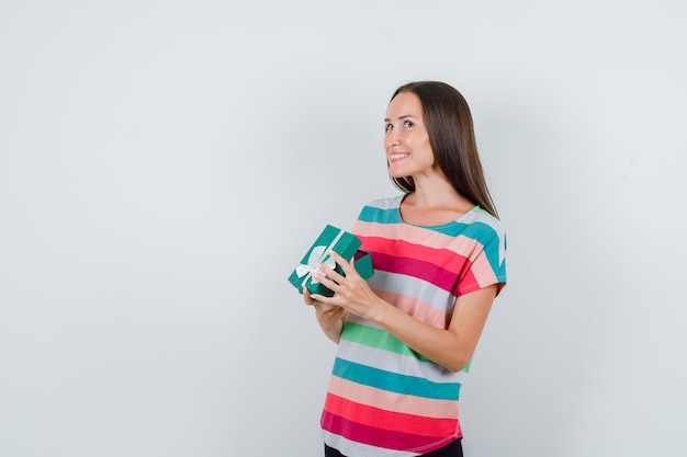 Mujer joven en camiseta, pantalones sosteniendo una caja de regalo abierta y mirando emocionado, vista frontal.