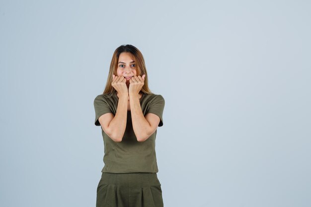 Mujer joven en camiseta, pantalones tomados de la mano cerca de la boca y mirando feliz, vista frontal.