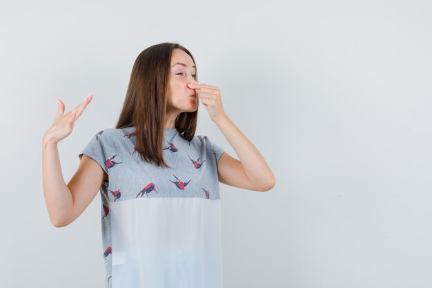 Mujer joven en camiseta pellizcando la nariz debido al mal olor y mirando disgustado, vista frontal.