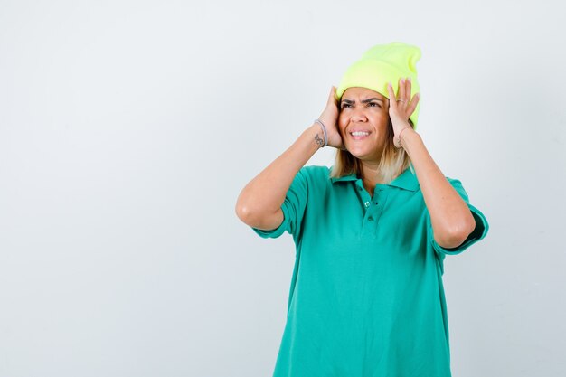 Mujer joven en camiseta de polo, gorro manteniendo las manos en la cabeza y mirando alegre, vista frontal.