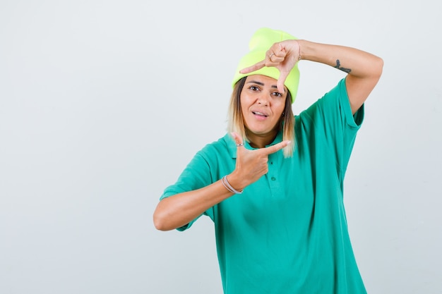 Mujer joven en camiseta de polo, gorro mostrando gesto de marco y mirando contento, vista frontal.