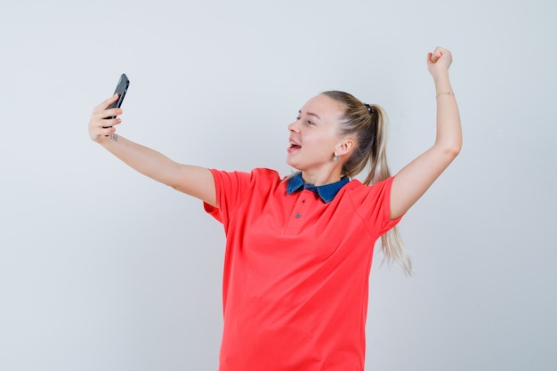 Mujer joven en camiseta que muestra el gesto del ganador mientras toma selfie y se ve juguetón