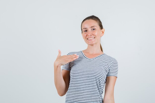 Mujer joven en camiseta a rayas apuntando a sí misma y mirando confiada, vista frontal.