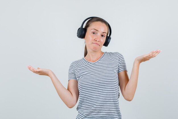 Mujer joven en camiseta a rayas, auriculares mostrando gesto de impotencia, vista frontal.