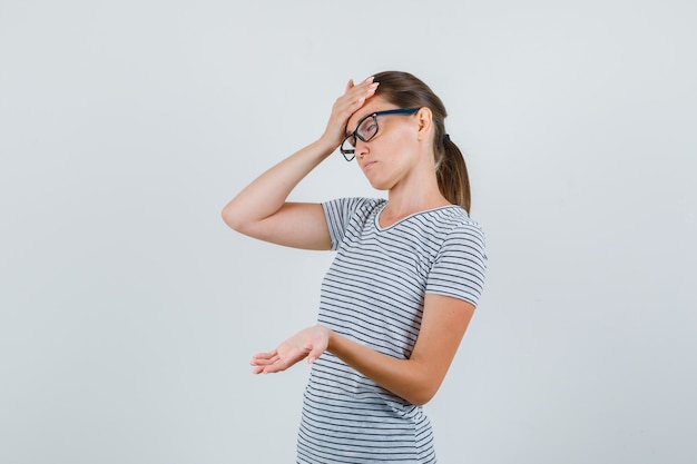 Mujer joven en camiseta a rayas, gafas mostrando la palma y con aspecto cansado, vista frontal.