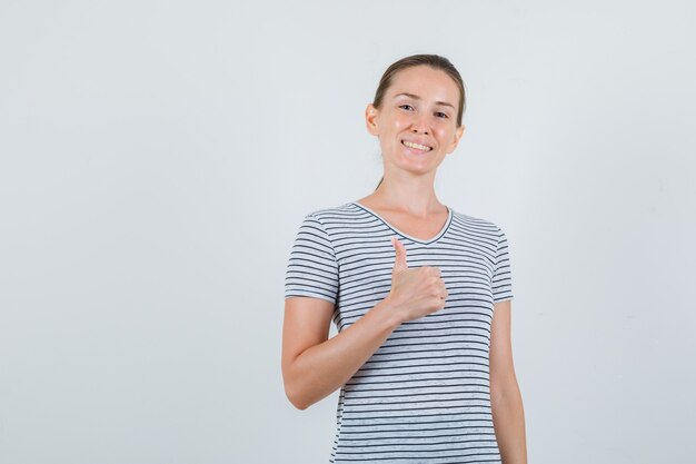 Mujer joven en camiseta de rayas mostrando el pulgar hacia arriba y mirando alegre, vista frontal.