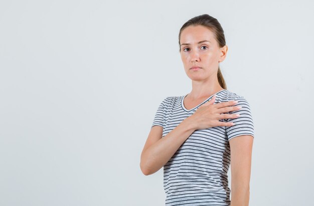 Mujer joven en camiseta a rayas sosteniendo la mano en el corazón y mirando seria, vista frontal.