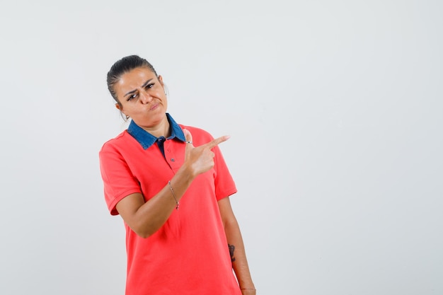 Foto gratuita mujer joven en camiseta roja apuntando a la derecha con el dedo índice y mirando disgustado, vista frontal.