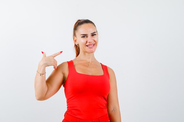 Mujer joven en camiseta roja apuntando hacia el lado derecho y mirando feliz, vista frontal.