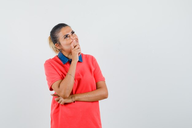 Mujer joven en camiseta roja con la mano apoyada en la boca, de pie en pose de pensamiento y mirando pensativo, vista frontal.