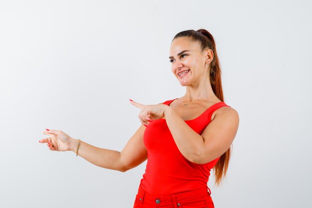 Mujer joven en camiseta roja, pantalones apuntando hacia el lado izquierdo y mirando feliz, vista frontal.