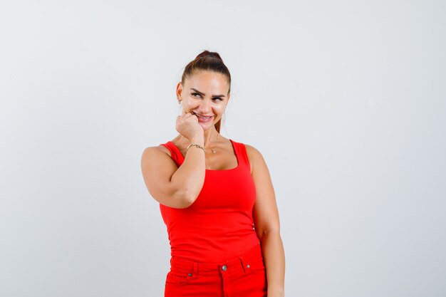 Mujer joven en camiseta roja, pantalones con la barbilla apoyada en el puño y luciendo atractiva, vista frontal.