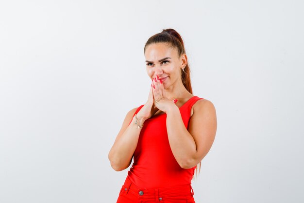 Mujer joven en camiseta roja, pantalones mostrando las manos juntas en gesto de súplica y mirando esperanzado, vista frontal.