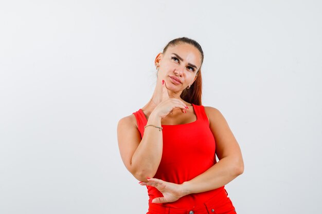 Mujer joven en camiseta roja, pantalones de pie en pose de pensamiento y mirando pensativo, vista frontal.