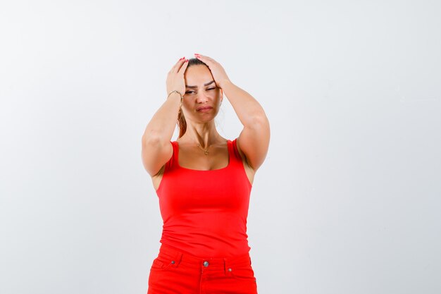 Mujer joven en camiseta roja, pantalones sosteniendo la cabeza con las manos y mirando molesto, vista frontal.