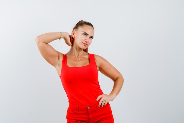 Mujer joven en camiseta roja, pantalones sosteniendo la mano en el cuello y la cintura y mirando pensativo, vista frontal.